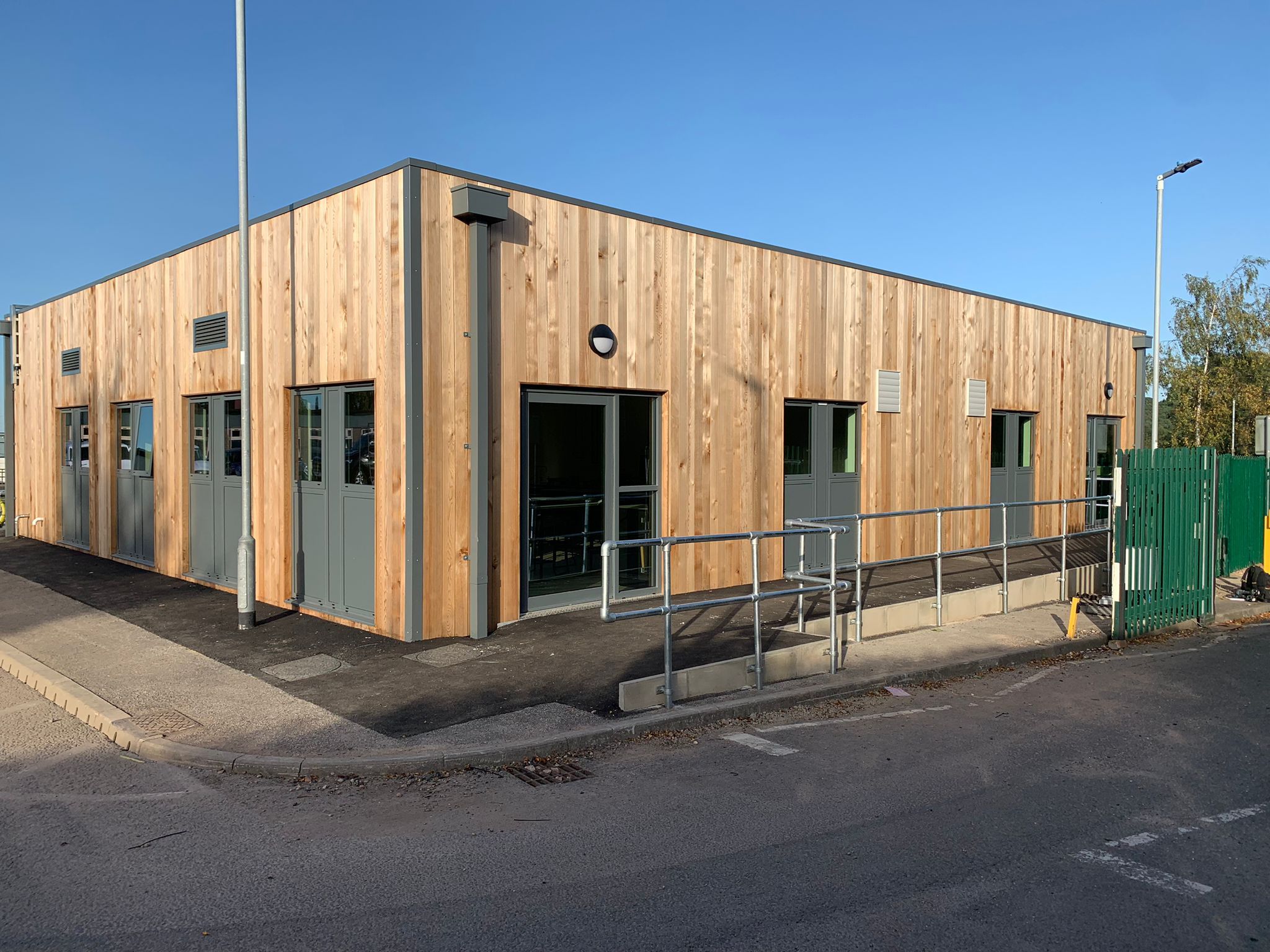 Wooden clad Modular Building. A rectangular, single-story wooden building features metal doors and windows, with a ramp and railing leading to the entrance. It sits adjacent to a street, under a clear blue sky.