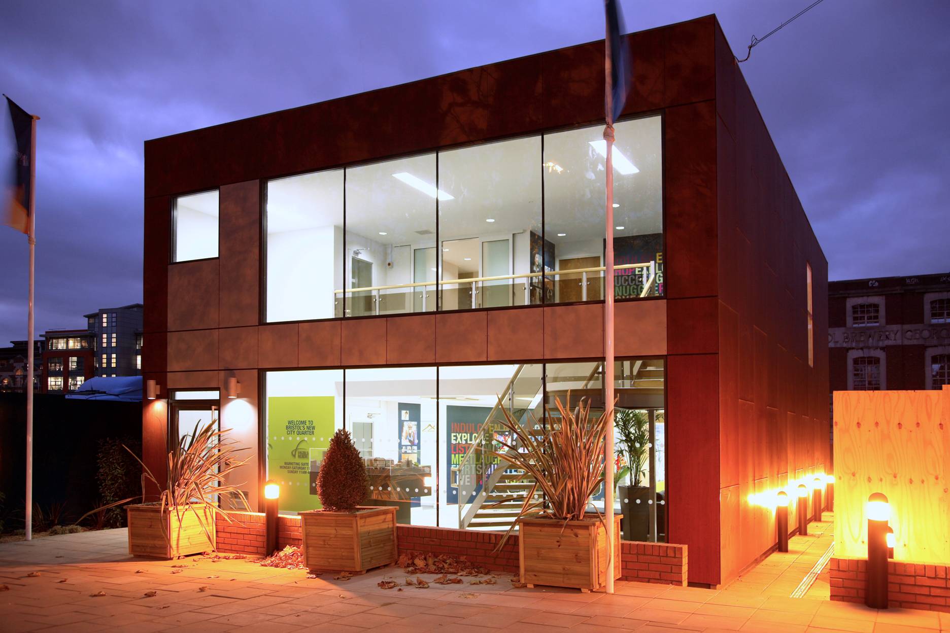 Modern, two-story building with large windows; indoor lights illuminate the interior, featuring stairs and potted plants; exterior includes planters and pathway lights; surrounded by a twilight urban environment. Text: &ldquo;Welcome to York House&rdquo; and other smaller print.