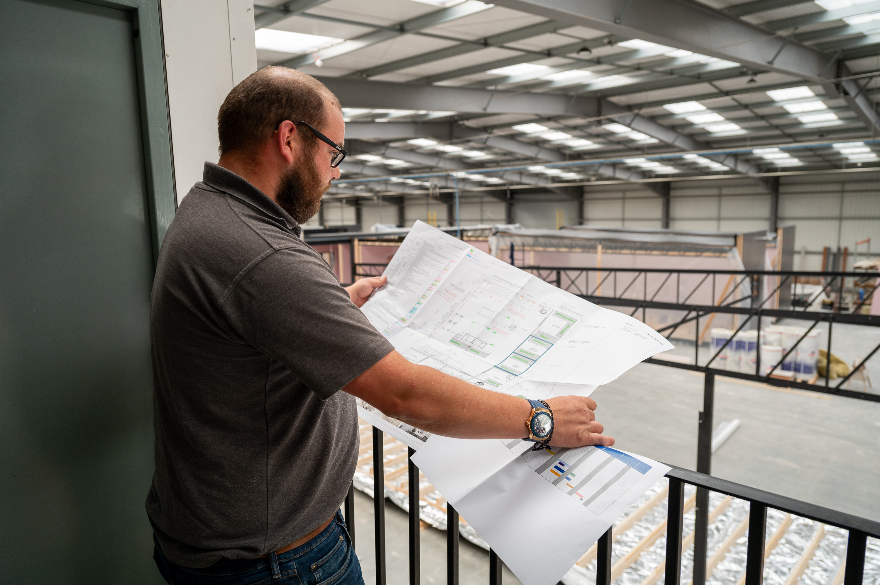 A man examines large architectural plans on an indoor mezzanine, overlooking a spacious, unfinished industrial warehouse with high ceilings, metal beams, and construction materials scattered throughout.