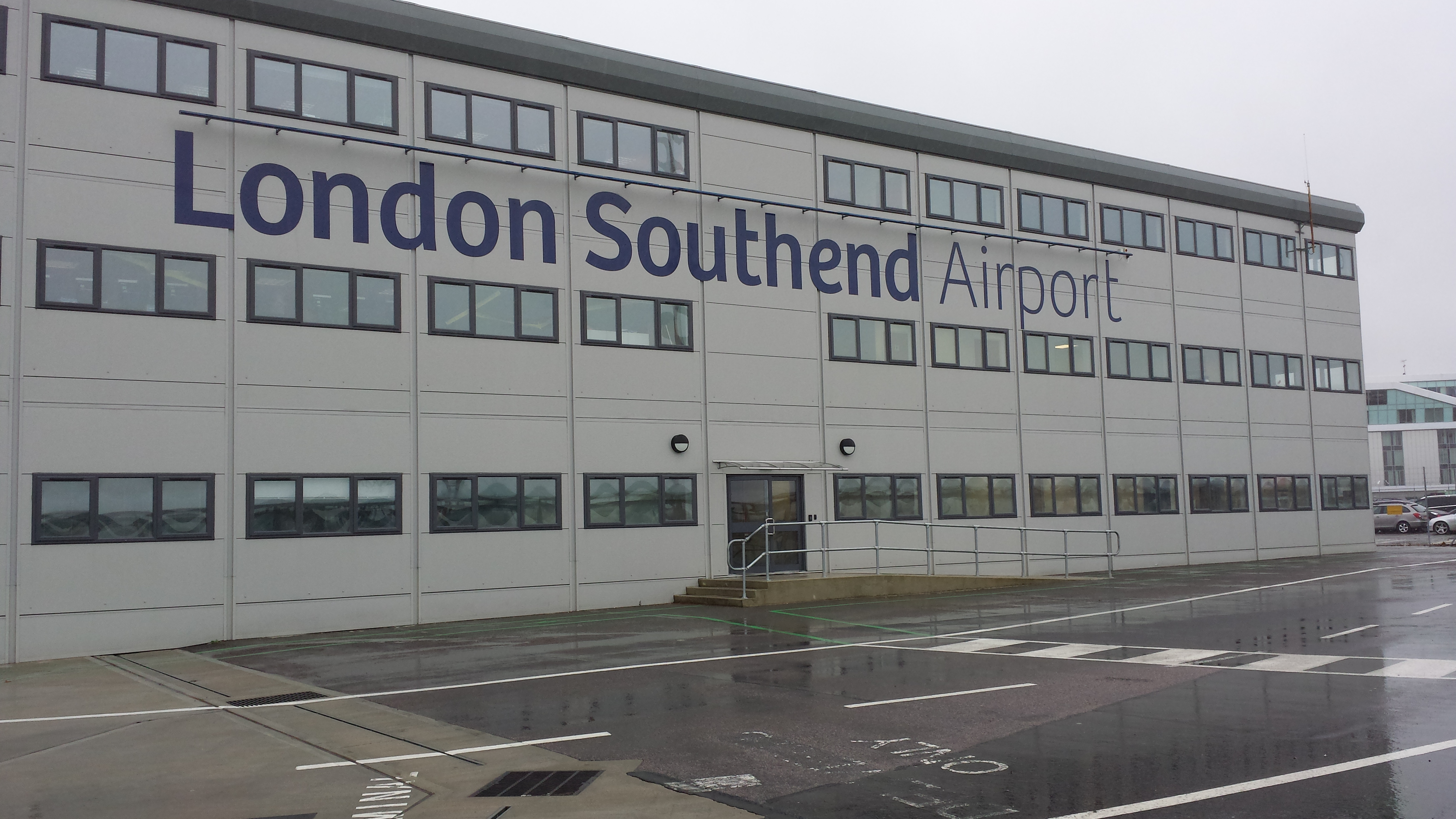 Building labeled 'London Southend Airport' stands in rainy weather, featuring multiple windows and a central entrance with a ramp. Surrounding area is a wet, empty parking lot.